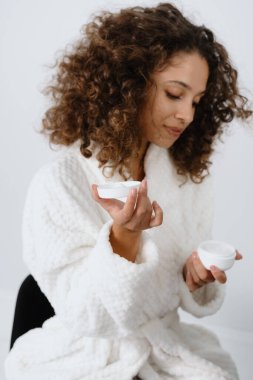 Evde spa prosedürü kavramı. Bornozlu, elinde nemlendirici krem olan, banyoda oturan ve rutin cilt bakımını yapan sakin Afro-Amerikan kadının dikey yan görüntüsü.