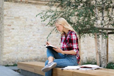 Odaklanmış, genç ve lise öğrencilerinin düşünceli yüz ifadeleri, ödev ödevleri hazırlama, kitaplara notlar yazma, kampüs binasının yanında oturma.
