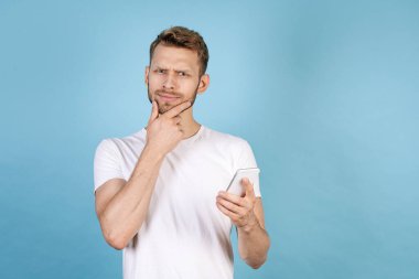 Modern akıllı telefonu elinde tutan düşünceli genç bir adam, gülen ve düşünen bir yüz yapıyor, mavi fotokopi arkaplanında tek başına duruyor, günlük beyaz tişört giyiyor.