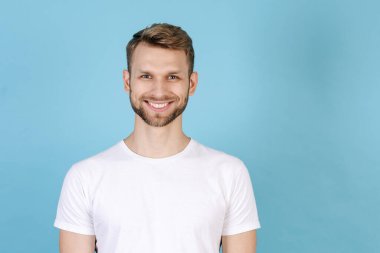 Beyaz tişört giyen mutlu genç bir adamın portresi, mavi fotokopi arkaplanında izole bir şekilde duruyor, kameraya bakıyor ve geniş bir gülümseme sergiliyor.