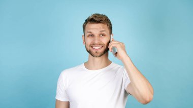 Modern akıllı telefondan konuşan mutlu ve genç bir adamın panoramik görüntüsü, geniş gülümseyen, mavi fotokopi arkaplanda beyaz tişörtle tek başına duran