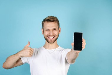 Reklam konsepti. Mutlu ve gülümseyen beyaz tişörtlü genç adam başparmaklarını kaldırıyor, elinde boş ekran ekranlı modern akıllı telefon tutuyor, mavi fotokopi arkaplanda tek başına duruyor.
