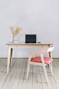 Modern iç tasarımın modern dairedeki dikey fotoğrafı ahşap masa, sandalye, akıllı telefonun yanındaki vazoda kuru bitkiler ve oturma odasındaki dizüstü bilgisayarın fotokopi duvarına karşı.