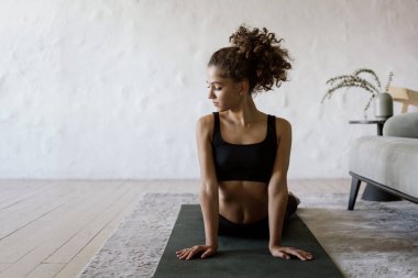 Sakin ve ciddi Afro-Amerikan kadın spor yapıyor. Evde yoga minderi üzerinde spor yapıyor. Spor kıyafetleri eğitimi alıyor, arka planda beyaz fotokopi duvarı var.