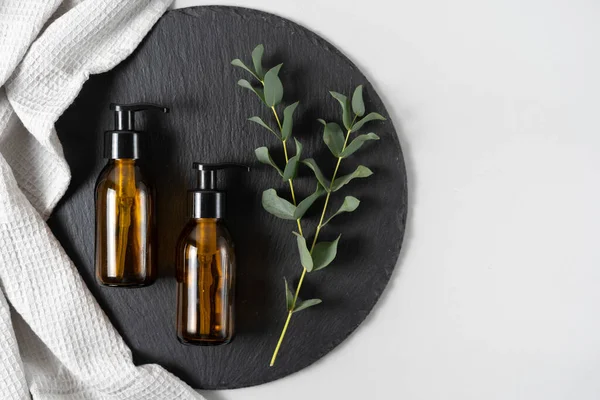 Zero waste, plastic free concept. Flat lay top view of natural and organic cosmetics in glass dispenser bottle near towel with eucalyptus plant on black plate stand against white copy space background