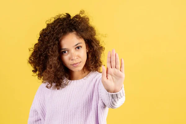 Stop violence and discrimination. Portrait of serious young afro american woman holding hand in front, making reject sign, standing on yellow copy space background