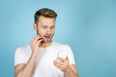 Şok olmuş genç bir adam modern akıllı telefon ekranına bakıyor, heyecanlı bir yüz ifadesi takınıyor, mavi fotokopi çekilen arka planda beyaz tişörtle dikiliyor. Çevrimiçi mağazada satış ve indirim kavramı