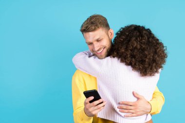 İnternet bağımlılığı konsepti. Mutlu genç adam Afro-Amerikan kadınına sarılıyor ve arkasında modern akıllı telefon kullanıyor. Mavi fotokopi arkaplanda tek başına duruyor.