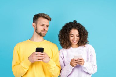 İlişki çatışması ve internet bağımlılığı kavramı. Ciddi bir yetişkin adam, Afro-Amerikan kadının internet uygulamasını kullanırken mavi arka planda tek başına duran akıllı telefon görüntüsüne bakıyor.
