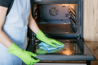 Parlak bir mutfakta, yeşil eldivenli bir kadın evdeki ev işleri sırasında temiz ve hijyenik bir yemek alanı sağlamak için fırın kapısını titizlikle temizler..