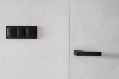 Bu modern iç mekan, siyah metal saplı kapıya yakın dokulu beyaz duvarlarda parlak minimalist bir ışık düğmesi sergiliyor ve konut alanındaki çağdaş ve şık bir tasarımı vurguluyor.