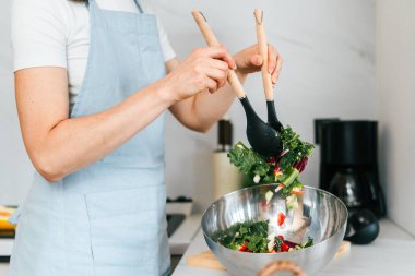 Bir kadın, parlak ve iyi donanımlı bir mutfakta önlük giyerken paslanmaz çelik bir kasede sebze salatası karıştırıyor. Sağlıklı bir yemek yapmaya odaklanmış..