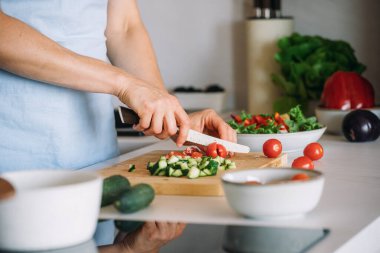 Kadın ahşap kesme tahtasına domates ve salatalık doğruyor. Kadın evde modern mutfakta salata pişiriyor. Bir kase taze sebze, sağlıklı bir yaşam tarzı.