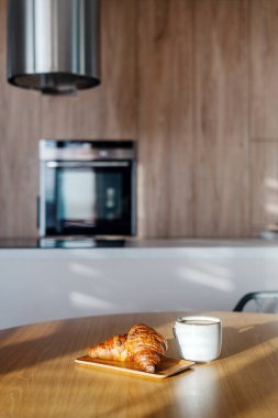 Sıcak, davetkar bir mutfak, taze kruvasanlarla dolu ahşap bir masa ve bir fincan sabah kahvesi, hafta sonu evde rahat bir an için mükemmel bir ortam yaratıyor.