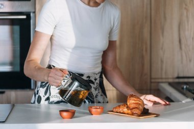 Bir insan küçük bardaklara buharlı çay dökerken altın kruvasan tabağı sıcak ve davetkar bir atmosferde mutfak tezgahında oturur.