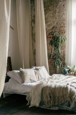 Kırsal yatak odasının içinde yumuşak çarşaflar ve dekoratif yastıklarla süslenmiş rahat bir yatak var. Doğal ışık perdelerin arasından süzülüp boho şık atmosferini güçlendirir..