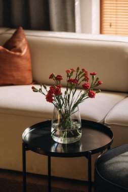 Modern bir oturma odasında şık beyaz bir kanepe ve parlak siyah bir kahve masası bulunur. Parlak kırmızı çiçeklerden oluşan bir vazo, sıcacık, davetkar yere renk katar..