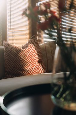 Oturma odasında, jaluzilerden süzülen sıcak güneş ışığı, koltuktaki dekoratif yastıklar ve yan masadaki basit çiçek aranjmanları sergileniyor..