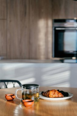Sıcak ve davetkar bir mutfak, içinde cam çaydanlık ve bir tabak kruvasan bulunan ahşap bir masa seti içerir. Sıcak bir dairede rahatlatıcı bir kahvaltı için mükemmeldir..