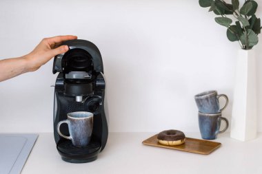 Bir el bir espresso makinesini açar ve içine kahve kapsülü koyar. Çağdaş bir mutfakta ev aletleri. Gri kupalar ve çörekler ahşap tepside oturur ve sabah rahat bir atmosfer yaratırlar..