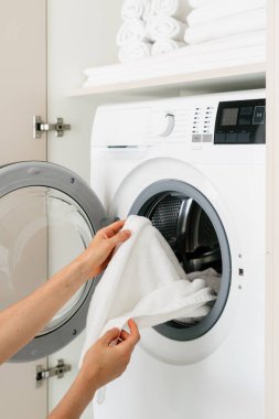 Parlak bir çamaşır odasındaki otomatik çamaşır makinesinden yeni yıkanmış beyaz havluları çıkartıyor. Mekanda modern cihazlar ve organize havlular, dikey çekim