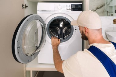 Otomatik çamaşır makinesinin içine bakıp davul ve prezervatifleri kontrol eden bir adam. Ev aletlerinin garanti hizmetleri. Çağdaş banyo, modern aletlerle dolu çamaşır odası.