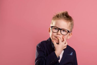 Okullu çocuk heyecanla tırnak yiyor. Bulling ve çocukluk psikolojisi, üniformalı çocuk, gözlük ve sırt çantası pembeye izole edilmiş. Parmağını ağzına sokmuş çocuk portresi.