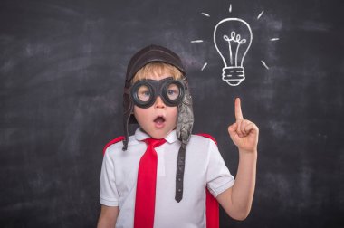 Okul çocuğu üniformalı, pilot kostümlü ve süper kahraman kırmızı pelerinli karatahtaya karşı oynuyor..