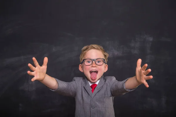 Funny school boy in uniform and glasses open arms toward camera and shows tongue. Happy child with surprised silly face against blackboard. Back to school. Education success winner innovation concept