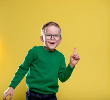 Sarı duvarda gözlüklü komik küçük çocuk. İlkokuldaki çocuğun bir fikri var. Sarı ceketli, yeşil kazaklı şık çocuk..