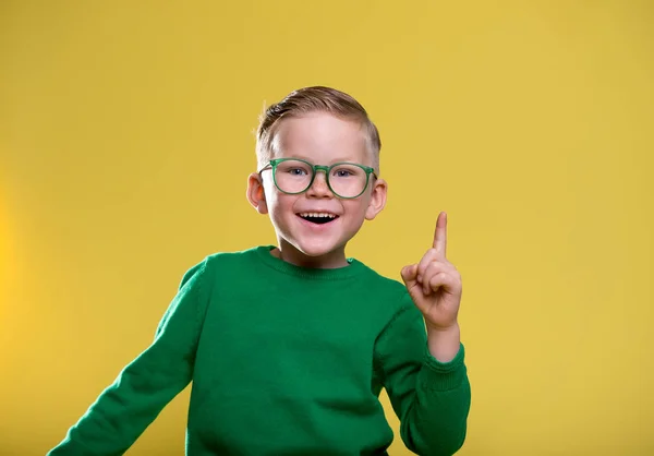 戴眼镜的有趣的小男孩指着黄色的墙壁 小学生有自己的想法 穿着黄色外套和绿色毛衣的时髦孩子 — 图库照片