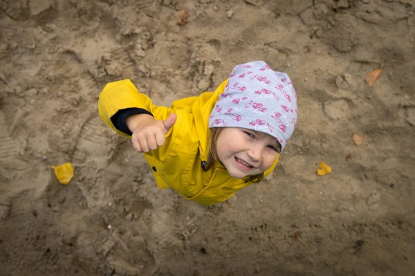Happy child in yellow coat playing in the sand box in playground. Kindergarten or daycare for kids. Autumn holiday. Happy smiling girl