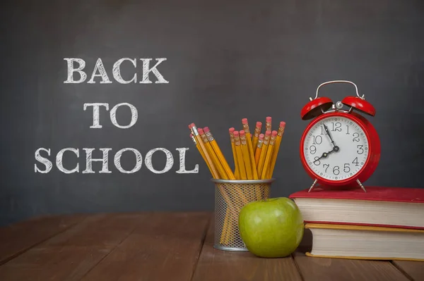 Desk, alarm clock, apple, pencils and school books against blackboard with back to school on background. Education.