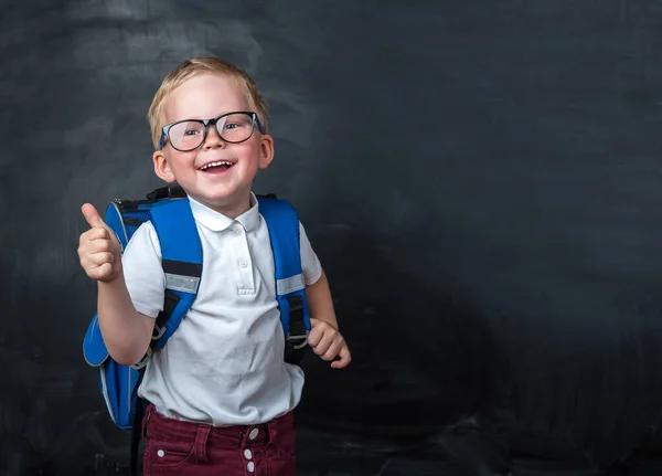 Happy smiling boy in glasses with thumb up is going to school for the first time. Child with school bag and book. Kid indoors of the class room with blackboard on a background. Back to school.