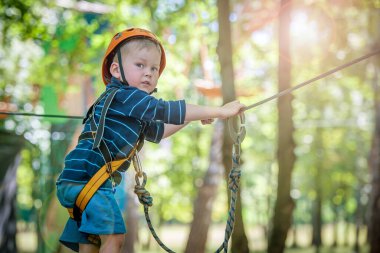 Küçük çocuk macera parkına kask ve güvenlik ekipmanıyla tırmanıyor..