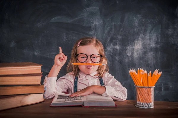Πίσω στο σχολείο. Αστείο χίπστερ κοριτσάκι στην αίθουσα του σχολείου που διασκεδάζει με μολύβι. Στοίβα από βιβλία στο γραφείο. Φίλτρο Instagram. Το παιδί δείχνει προς τα πάνω. Λαμπρή ιδέα. — Φωτογραφία Αρχείου