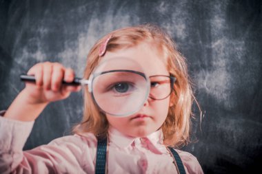 Küçük kız büyüteçle tahtaya bakıyor. Instagram filtresi. Okula dönelim. Komik çocuk suratı. Gözbebeği.