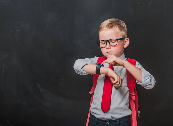 Little kid boy in glasses and school uniform is looking on smart watch on his hand against blackboard background. Back to school concept.