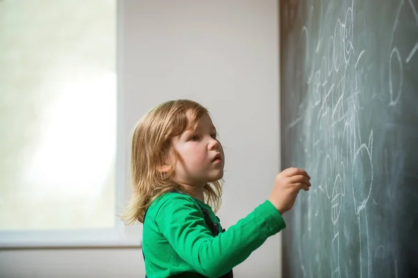Child girl writing on chalkboard. Back to school. Kid with chalk in hand in class. Thinking pupil. Little genius.