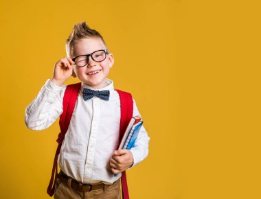 Okuldaki ilk gününde gözlüklü, kitaplı ve çantalı mutlu bir çocuk. Sarı arka plana karşı komik çocuk. Eğitim. Okul konseptine geri dönelim. Dahi..