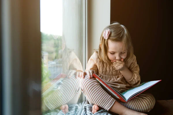 어린 소녀는 창문 가까이에서 책을 읽었다. 교육과 학교의 개념. 책을 손에 쥐고 있는아이. 빠르게 읽기 — 스톡 사진