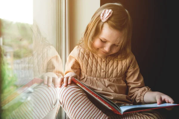 어린 소녀는 창문 가까이에서 책을 읽었다. 교육과 학교의 개념. 책을 손에 쥐고 있는아이. 빨리 읽어. 천재 — 스톡 사진