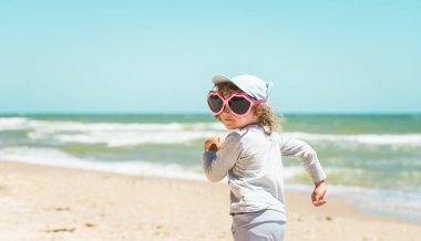 Sahilde büyük güneş gözlüklü komik küçük kız. Yaz tatili ve aşk. Kırmızı dudak gibi güneş gözlüğü. Sahildeki mutlu çocuk. Şık koşan çocuk.