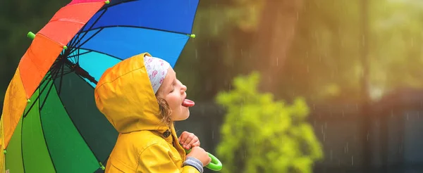Happy and free little girl opens moth and enjoys of rain. Little girl hiding under an umbrella from the rain. Happy childhood concept. Kid catching rain drops with tongue. Bright style clothes.