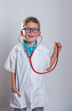 Steteskopla doktorculuk oynayan bir çocuk. Gözlüklü çocuk beyazdan soyutlanmış. Başparmak yukarı. Beyaz elbiseler.