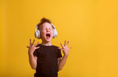 Beyaz kulaklıkla müzik dinlerken çocuk yüksek sesle şarkı söylüyor. Şık çocuk sarıda izole edilmiş. Elleri duygusal olarak açık bir çocuk. Müzik notaları resmi.