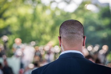 Security guard listening to his earpiece on event. Back of jacket showing. secret service guard. private bodyguard. man with earpiece in crowd. Black suit and bald head. clipart