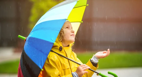 Funny little girl playing in the garden under the autumn rain. Kid wearing yellow waterproof coat and boots holding colorful umbrella. Rainy weather. Child is catching drops — Stock Photo, Image