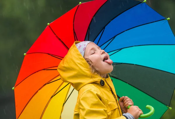 Забавная маленькая девочка играет в саду под осенним дождем. Парень в жёлтом водонепроницаемом пальто и ботинках с красочным зонтиком. Дождливая погода. Ребенок ловит капли. Семейная прогулка в парке. — стоковое фото