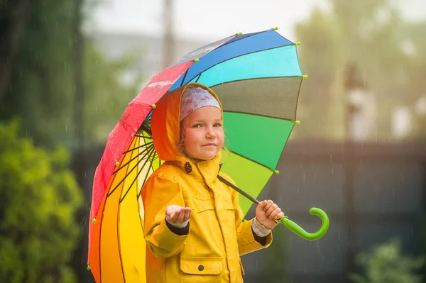 Маленькая девочка с зонтиком под дождем. Ребенок в водонепроницаемом желтом пальто и резиновых сапогах. Ребенок веселится в дождливый день — стоковое фото